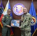Two military officers are posing together in front of a Coast Guard Aviation Command seal and multiple organizational flags. They are jointly holding a framed commemorative plaque or shadow box, suggesting an official exchange or ceremony. One officer is wearing a Philippine Coast Guard flight suit and the other is in U.S. Army multicam (OCP) uniform.