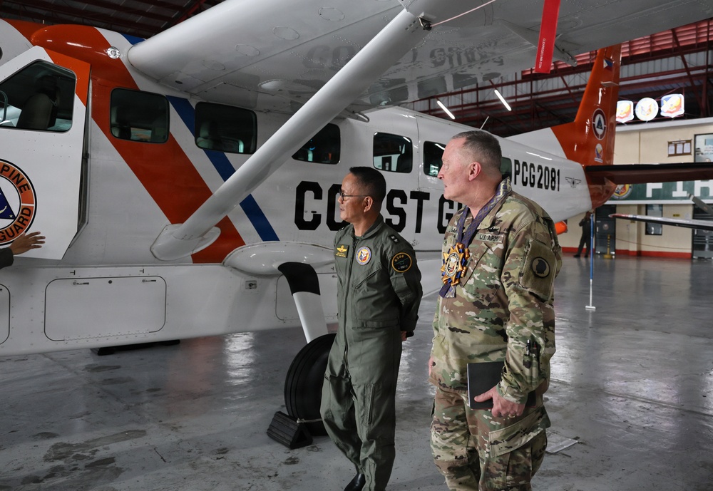 Two military personnel stand in an aircraft hangar in front of a Philippine Coast Guard aircraft marked 'COAST GUARD' and 'PCG 2081'. One individual wears an olive green flight suit with patches, while the other wears a US Army multicam combat uniform with a medal/decoration around his neck. They appear to be engaged in a briefing or tour of the facility.