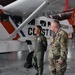 Two military personnel stand in an aircraft hangar in front of a Philippine Coast Guard aircraft marked 'COAST GUARD' and 'PCG 2081'. One individual wears an olive green flight suit with patches, while the other wears a US Army multicam combat uniform with a medal/decoration around his neck. They appear to be engaged in a briefing or tour of the facility.