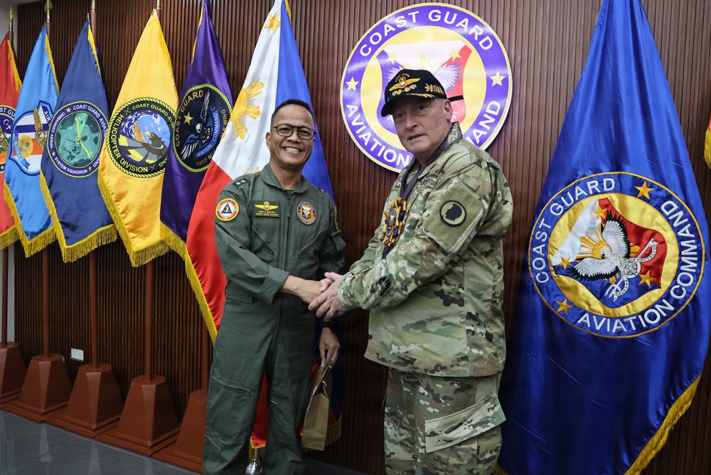 Two military officers are shaking hands in front of a display of organizational flags and a prominent 'Coast Guard Aviation Command' seal on a wooden wall. One officer is wearing an olive/green flight suit with patches, and the other is in a multicam/OCP camouflage uniform with a black cap. The setting appears to be an official meeting or visit at a Coast Guard Aviation Command headquarters.