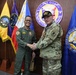 Two military officers are shaking hands in front of a display of organizational flags and a prominent 'Coast Guard Aviation Command' seal on a wooden wall. One officer is wearing an olive/green flight suit with patches, and the other is in a multicam/OCP camouflage uniform with a black cap. The setting appears to be an official meeting or visit at a Coast Guard Aviation Command headquarters.