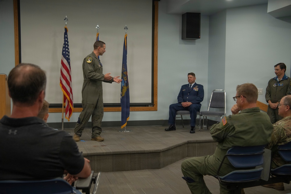 A military briefing or ceremony is taking place in an indoor meeting room. A person in an olive green flight suit is speaking or presenting on a raised platform stage, while an officer in a blue dress uniform sits behind them and an audience of military personnel in various uniforms observes. Two flags — the United States flag and what appears to be a U.S. Air Force flag — are displayed on the stage.