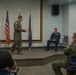 A military briefing or ceremony is taking place in an indoor meeting room. A person in an olive green flight suit is speaking or presenting on a raised platform stage, while an officer in a blue dress uniform sits behind them and an audience of military personnel in various uniforms observes. Two flags — the United States flag and what appears to be a U.S. Air Force flag — are displayed on the stage.