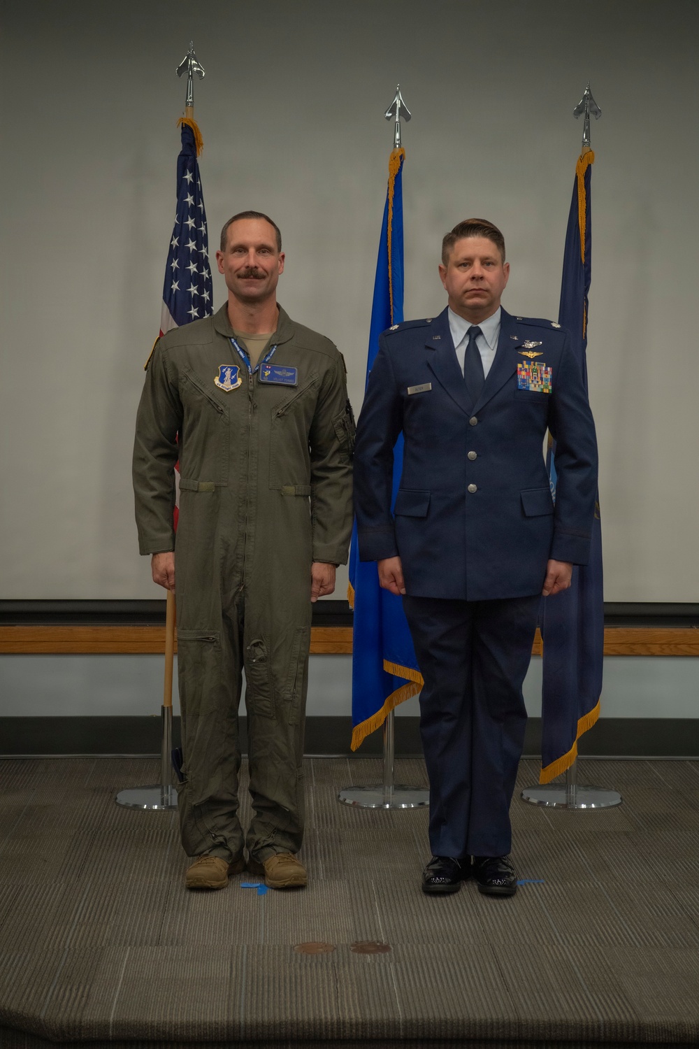 Two U.S. Air Force personnel are standing at attention in a formal military setting, likely during a ceremony or official photo. One individual is wearing an olive green flight suit with patches and pilot wings, while the other is in the Air Force Service Dress uniform with multiple ribbons and badges. Three flags are displayed behind them, including the U.S. flag and Air Force flags.