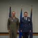 Two U.S. Air Force personnel are standing at attention in a formal military setting, likely during a ceremony or official photo. One individual is wearing an olive green flight suit with patches and pilot wings, while the other is in the Air Force Service Dress uniform with multiple ribbons and badges. Three flags are displayed behind them, including the U.S. flag and Air Force flags.