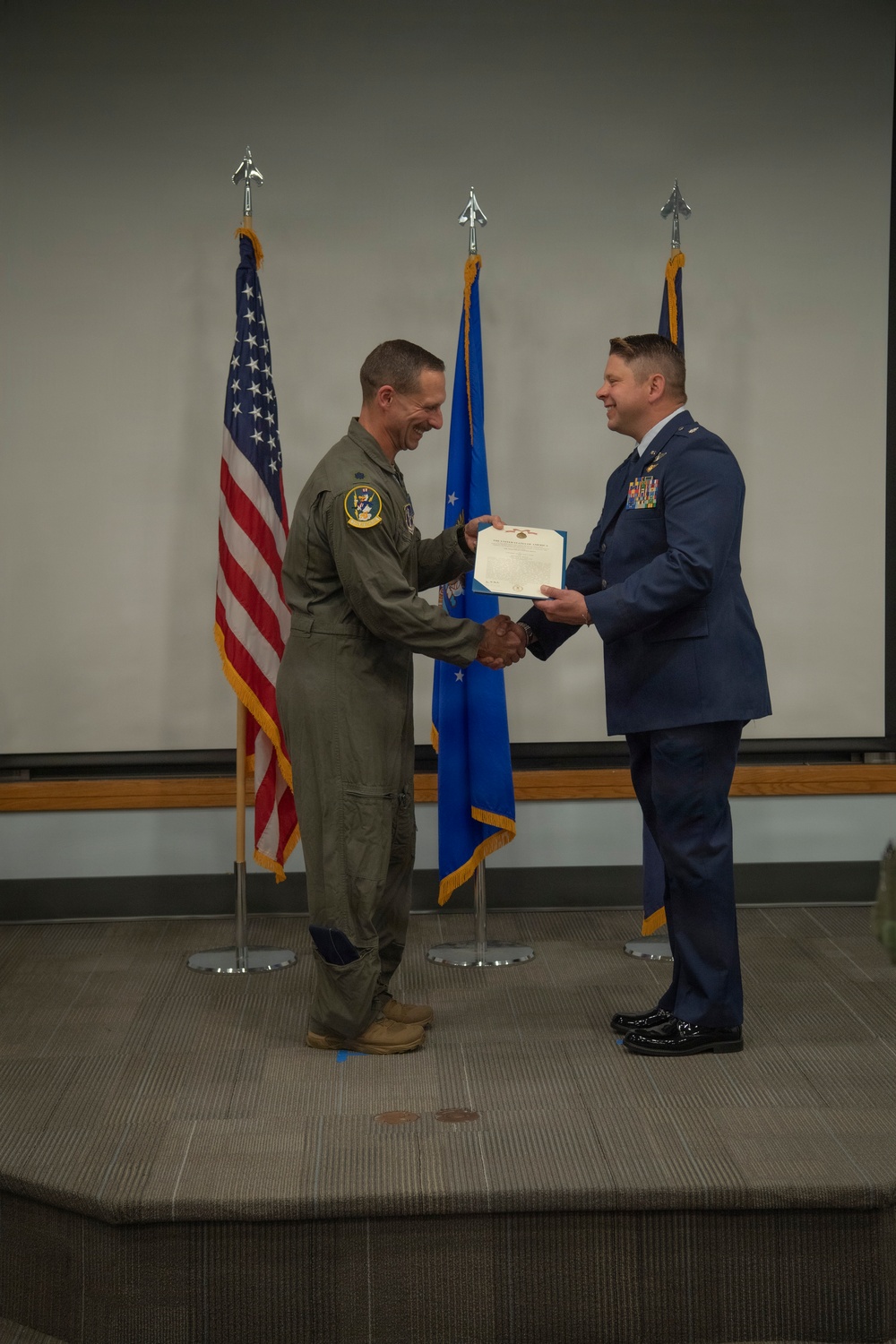 Two U.S. Air Force personnel are on a stage engaged in a formal military award or recognition ceremony. One individual is wearing an olive green flight suit with a squadron patch, while the other is in the blue Air Force service dress uniform with multiple ribbons and medals. They are shaking hands while jointly holding a certificate or award document.