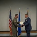Two U.S. Air Force personnel are on a stage engaged in a formal military award or recognition ceremony. One individual is wearing an olive green flight suit with a squadron patch, while the other is in the blue Air Force service dress uniform with multiple ribbons and medals. They are shaking hands while jointly holding a certificate or award document.