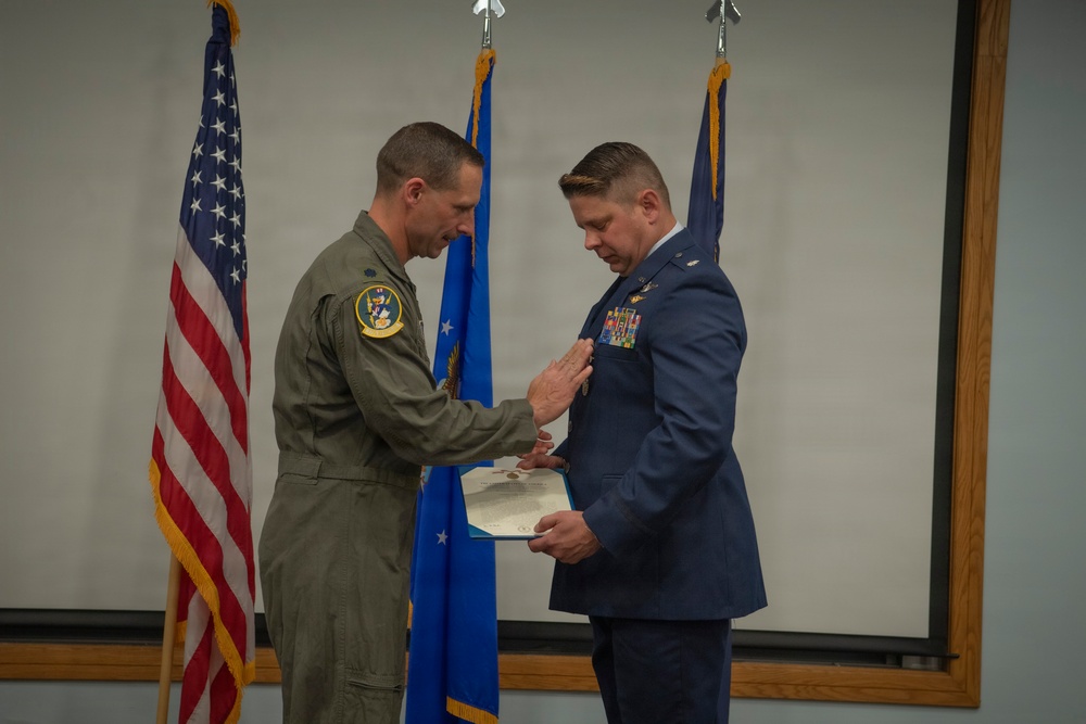 A military ceremony is taking place in which one officer in an olive green flight suit is pinning a medal or award onto another officer dressed in a formal US Air Force blue service uniform. The recipient is holding a certificate or citation document while the decoration is being applied to his chest. American and US Air Force flags are visible in the background.