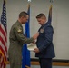 A military ceremony is taking place in which one officer in an olive green flight suit is pinning a medal or award onto another officer dressed in a formal US Air Force blue service uniform. The recipient is holding a certificate or citation document while the decoration is being applied to his chest. American and US Air Force flags are visible in the background.