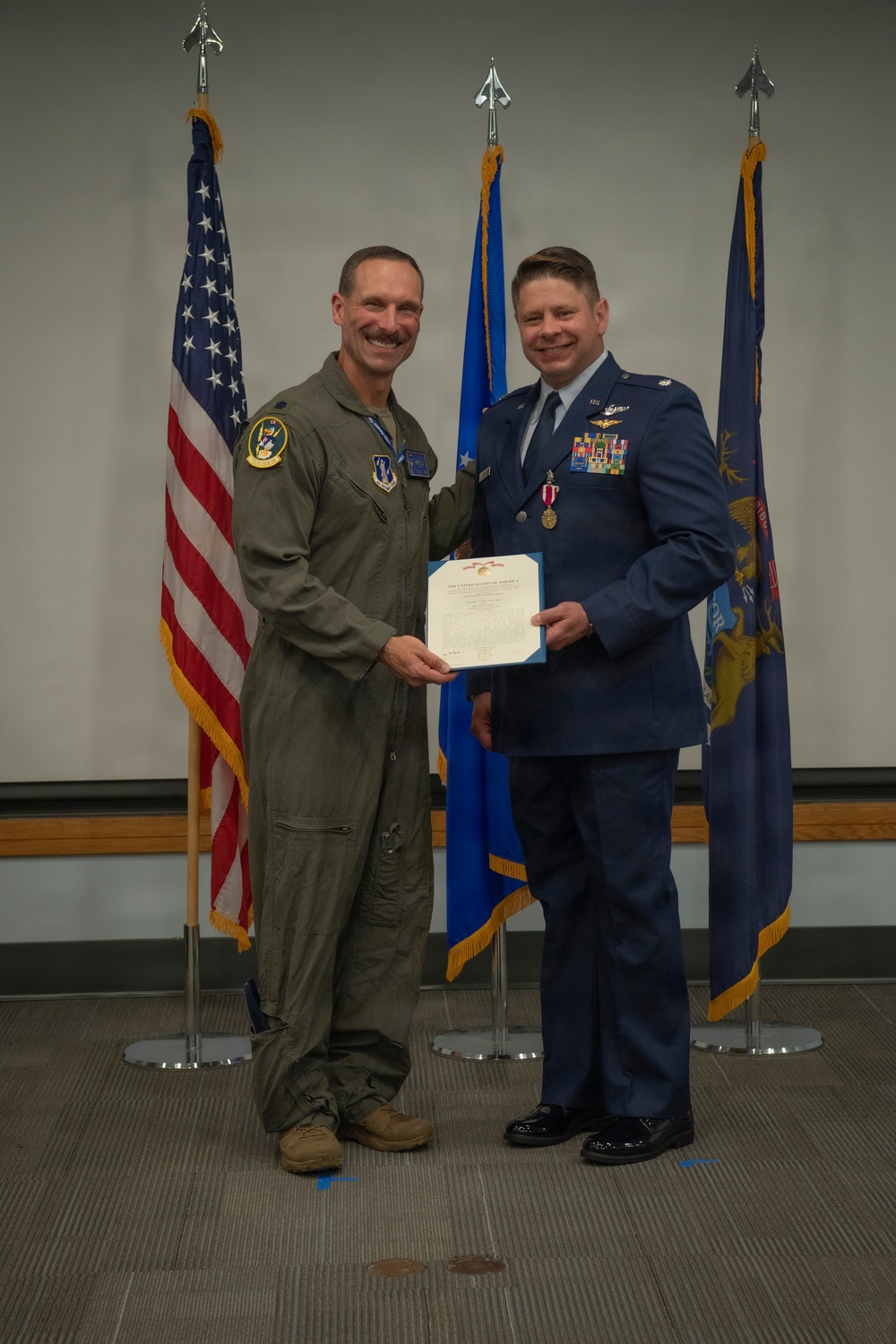 Two U.S. Air Force personnel pose together during a formal military awards ceremony. One individual wearing a green flight suit is presenting a certificate to another individual in a dress blue uniform who is also wearing a medal. Three flags are displayed in the background: the U.S. flag, an Air Force flag, and what appears to be a state or unit flag.