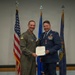 Two U.S. Air Force personnel pose together during a formal military awards ceremony. One individual wearing a green flight suit is presenting a certificate to another individual in a dress blue uniform who is also wearing a medal. Three flags are displayed in the background: the U.S. flag, an Air Force flag, and what appears to be a state or unit flag.