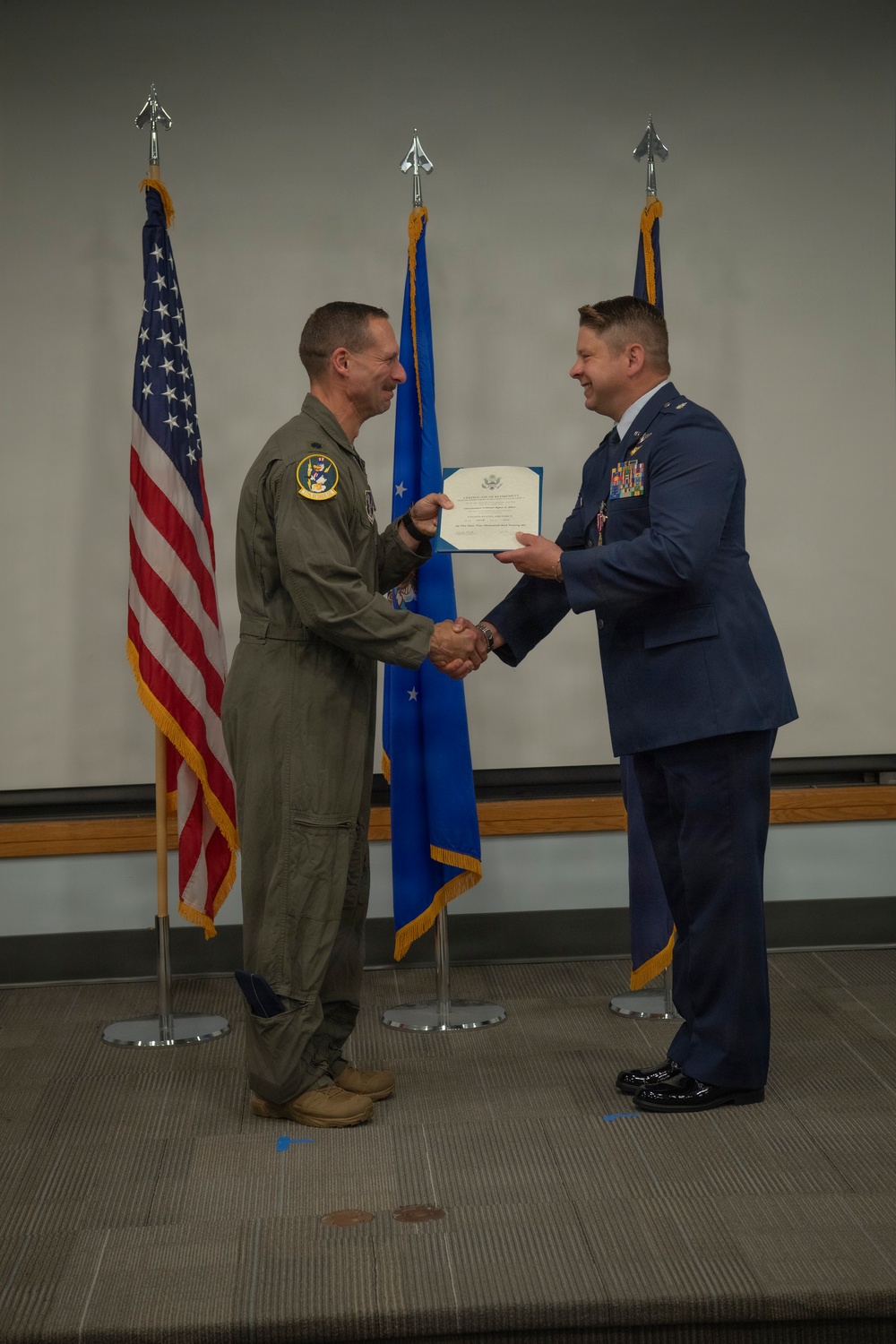 Two U.S. Air Force personnel are shaking hands during a formal military ceremony, with one presenting an official certificate or award document to the other. The individual on the left is wearing an olive green flight suit with a squadron patch, while the individual on the right is in a dress blue uniform adorned with multiple ribbons and medals. Three flags — the U.S. flag, an Air Force flag, and a unit flag — are displayed in the background.