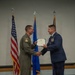 Two U.S. Air Force personnel are shaking hands during a formal military ceremony, with one presenting an official certificate or award document to the other. The individual on the left is wearing an olive green flight suit with a squadron patch, while the individual on the right is in a dress blue uniform adorned with multiple ribbons and medals. Three flags — the U.S. flag, an Air Force flag, and a unit flag — are displayed in the background.