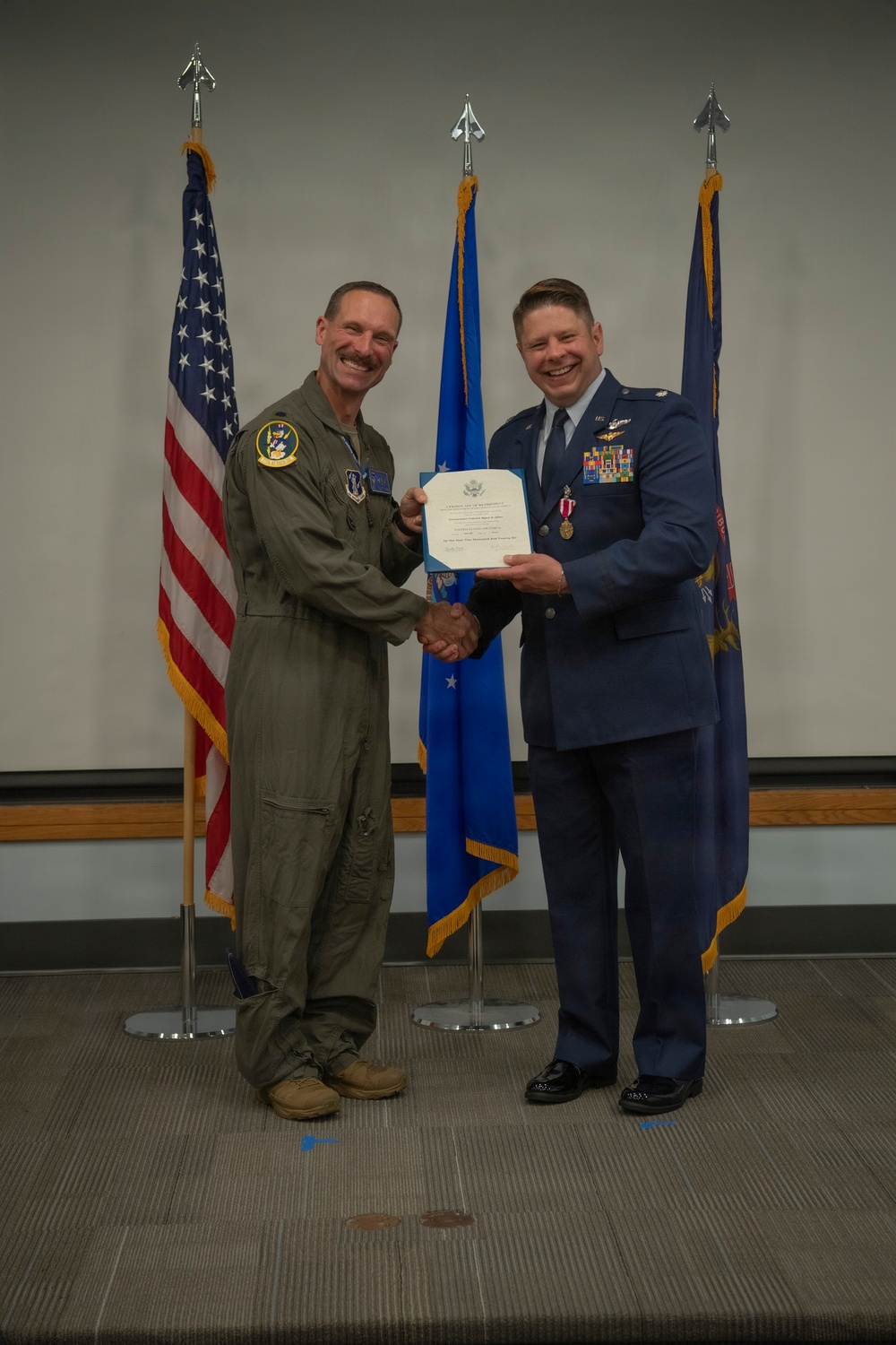Two U.S. Air Force personnel are posing for a formal ceremony photo, shaking hands while jointly holding an official certificate or award document. One individual is wearing an olive green flight suit with squadron patches, while the other is in the formal blue Air Force dress uniform adorned with medals and ribbons. Three flags are visible in the background: the U.S. flag, an Air Force flag, and what appears to be a unit or command flag.