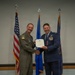 Two U.S. Air Force personnel are posing for a formal ceremony photo, shaking hands while jointly holding an official certificate or award document. One individual is wearing an olive green flight suit with squadron patches, while the other is in the formal blue Air Force dress uniform adorned with medals and ribbons. Three flags are visible in the background: the U.S. flag, an Air Force flag, and what appears to be a unit or command flag.