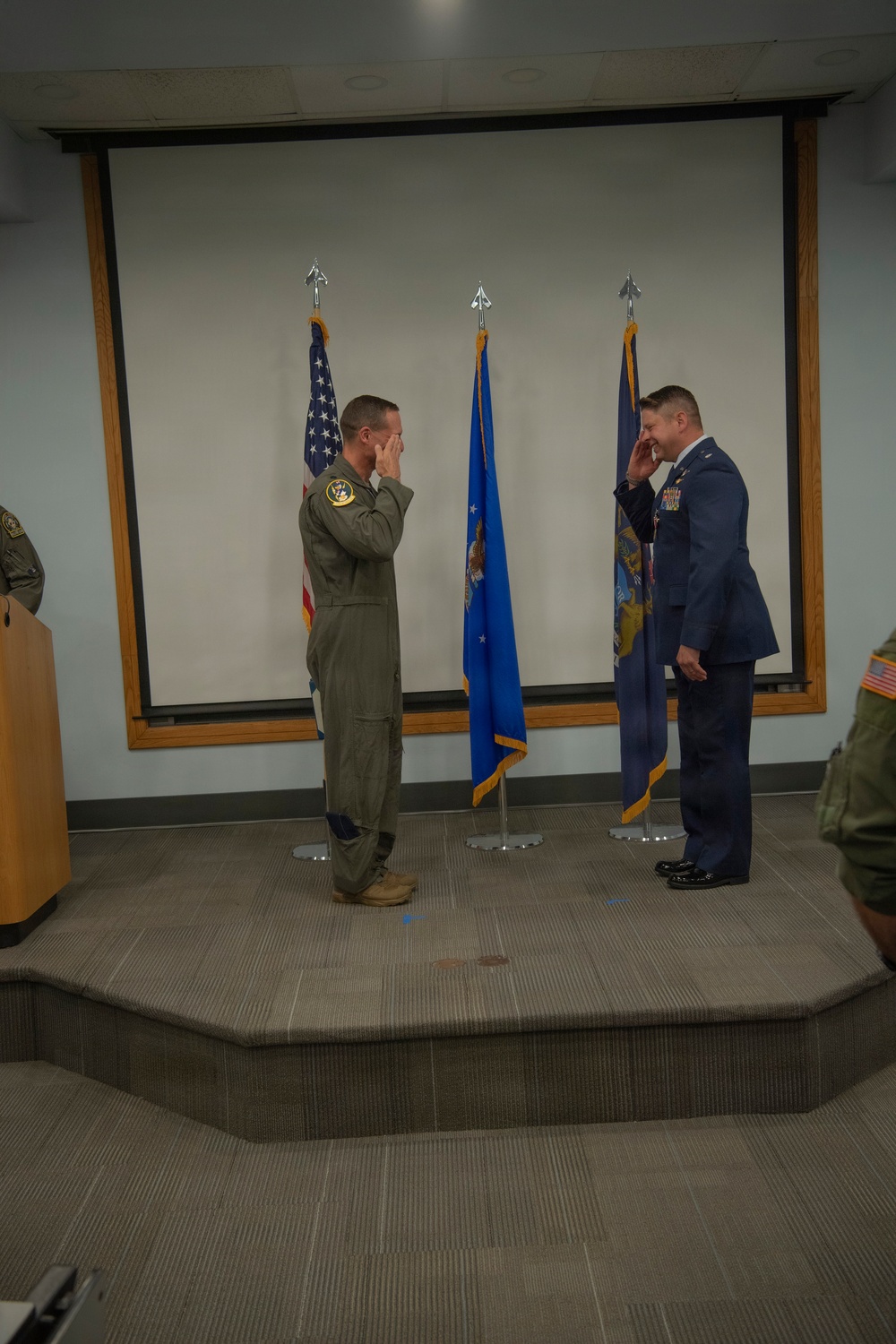 Two military personnel are exchanging salutes on a raised platform during what appears to be a formal ceremony. One individual is wearing an olive green flight suit with a squadron patch, while the other is in a formal blue Air Force dress uniform with medals and ribbons. Both men appear to be smiling or laughing during the salute exchange.