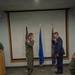 Two military personnel are exchanging salutes on a raised platform during what appears to be a formal ceremony. One individual is wearing an olive green flight suit with a squadron patch, while the other is in a formal blue Air Force dress uniform with medals and ribbons. Both men appear to be smiling or laughing during the salute exchange.