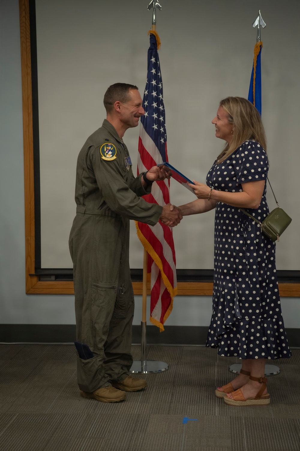 A military aviator in an olive green flight suit is shaking hands with a civilian woman in a navy blue polka dot dress while exchanging what appears to be a certificate or award folder. The ceremony is taking place indoors in front of a United States flag and what appears to be a light blue military branch flag. Both individuals are smiling, suggesting a positive recognition or award ceremony.