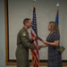 A military aviator in an olive green flight suit is shaking hands with a civilian woman in a navy blue polka dot dress while exchanging what appears to be a certificate or award folder. The ceremony is taking place indoors in front of a United States flag and what appears to be a light blue military branch flag. Both individuals are smiling, suggesting a positive recognition or award ceremony.