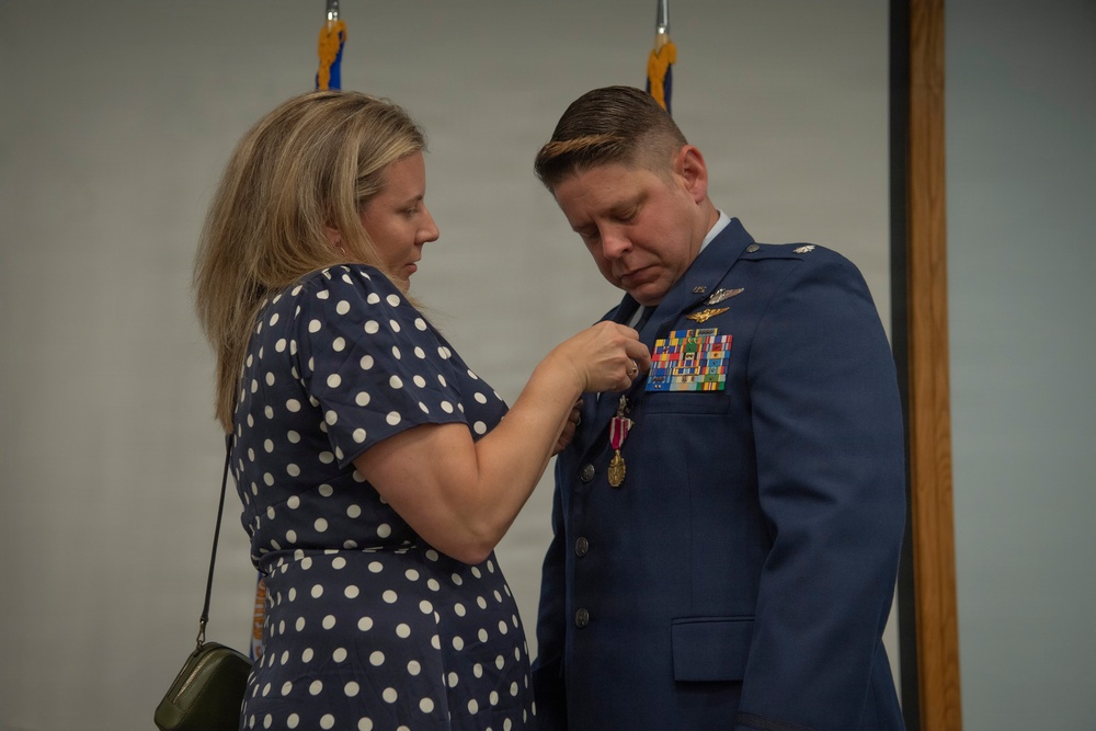 A woman in a navy blue polka-dot dress is pinning a medal onto the uniform of a male U.S. Air Force officer during what appears to be a formal military ceremony. The officer is wearing a dress blue uniform adorned with multiple ribbons, badges, and decorations. The setting appears to be an indoor military venue with flags visible in the background.