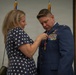 A woman in a navy blue polka-dot dress is pinning a medal onto the uniform of a male U.S. Air Force officer during what appears to be a formal military ceremony. The officer is wearing a dress blue uniform adorned with multiple ribbons, badges, and decorations. The setting appears to be an indoor military venue with flags visible in the background.