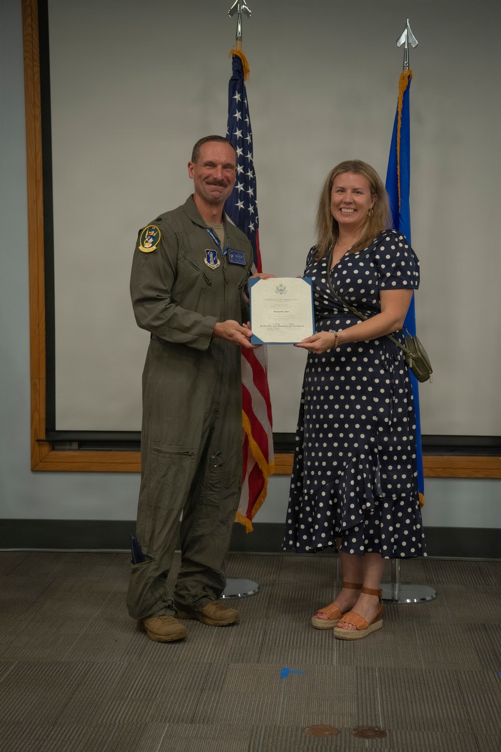A military officer in an olive green flight suit stands alongside a civilian woman in a navy blue polka-dot dress, both holding a certificate or award document. They are posed in front of an American flag and a U.S. Air Force flag, suggesting a formal recognition or award ceremony. The setting appears to be an indoor military facility or conference room.