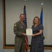 A military officer in an olive green flight suit stands alongside a civilian woman in a navy blue polka-dot dress, both holding a certificate or award document. They are posed in front of an American flag and a U.S. Air Force flag, suggesting a formal recognition or award ceremony. The setting appears to be an indoor military facility or conference room.
