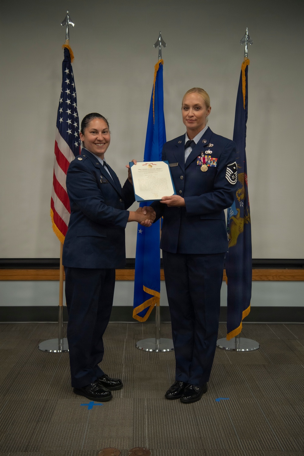 Two female United States Air Force personnel are posing together during a formal military ceremony, jointly holding an official certificate or award document. The individual on the right, a Senior Master Sergeant, is receiving recognition while shaking hands with an officer on the left. Three flags are displayed in the background: the U.S. flag, an Air Force flag, and what appears to be a state flag.