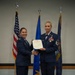 Two female United States Air Force personnel are posing together during a formal military ceremony, jointly holding an official certificate or award document. The individual on the right, a Senior Master Sergeant, is receiving recognition while shaking hands with an officer on the left. Three flags are displayed in the background: the U.S. flag, an Air Force flag, and what appears to be a state flag.