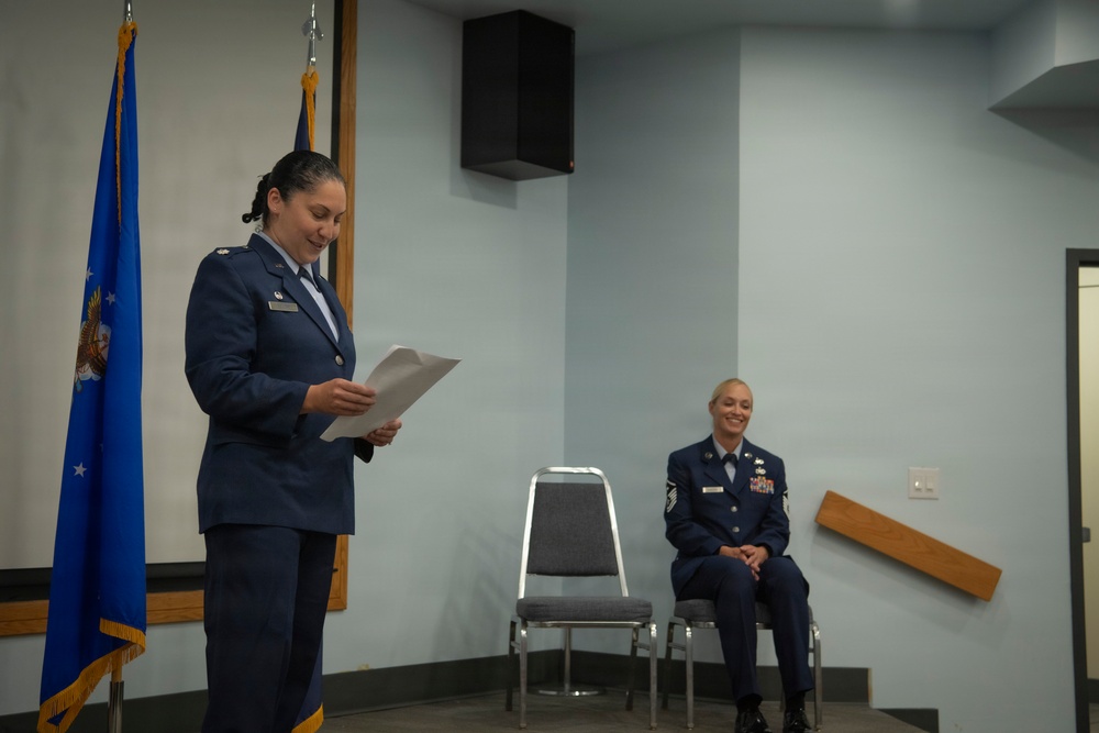 Two female U.S. Air Force personnel are participating in what appears to be a military ceremony or formal event indoors. One officer is standing at the front reading from a document, while a senior non-commissioned officer (NCO) is seated and smiling in the background. Both are wearing the Air Force Service Dress uniform (blue).