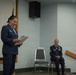 Two female U.S. Air Force personnel are participating in what appears to be a military ceremony or formal event indoors. One officer is standing at the front reading from a document, while a senior non-commissioned officer (NCO) is seated and smiling in the background. Both are wearing the Air Force Service Dress uniform (blue).