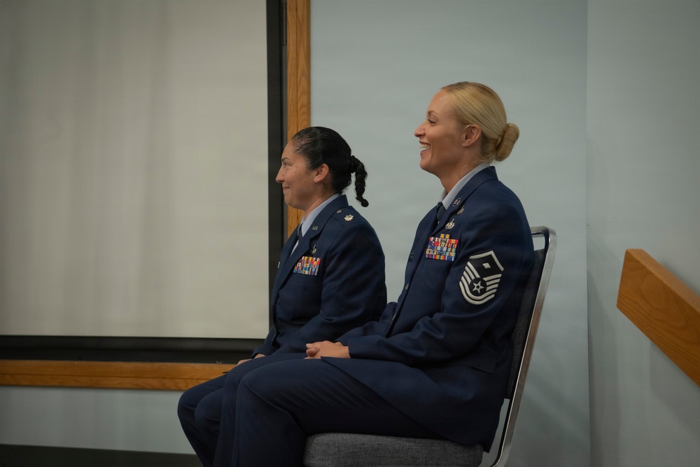 Two female U.S. Air Force personnel are seated side by side in what appears to be an indoor military ceremony or briefing room. Both are wearing the Air Force Service Dress uniform (blue) with visible rank insignia and service ribbons. The woman on the right is smiling broadly, while the woman on the left has a subtle smile.