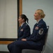 Two female U.S. Air Force personnel are seated side by side in what appears to be an indoor military ceremony or briefing room. Both are wearing the Air Force Service Dress uniform (blue) with visible rank insignia and service ribbons. The woman on the right is smiling broadly, while the woman on the left has a subtle smile.