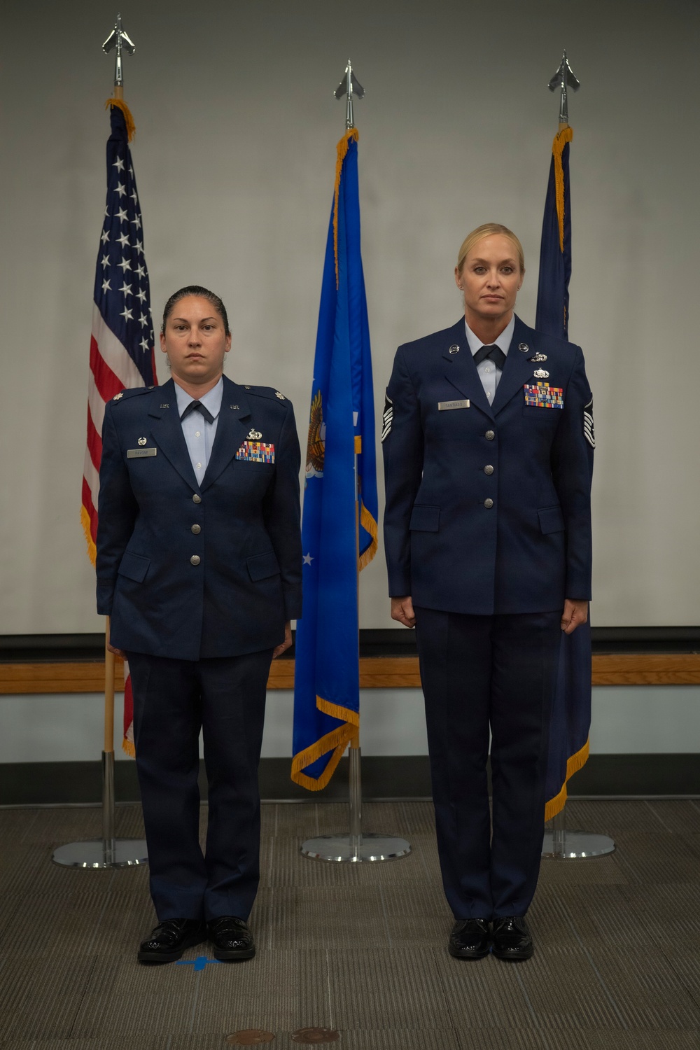 Two female United States Air Force personnel stand at attention in dress blue uniforms during what appears to be a formal military ceremony. They are positioned in front of three flags: the American flag, a blue Air Force flag, and a dark blue organizational flag. Both individuals display multiple service ribbons and badges on their uniforms.