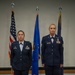 Two female United States Air Force personnel stand at attention in dress blue uniforms during what appears to be a formal military ceremony. They are positioned in front of three flags: the American flag, a blue Air Force flag, and a dark blue organizational flag. Both individuals display multiple service ribbons and badges on their uniforms.