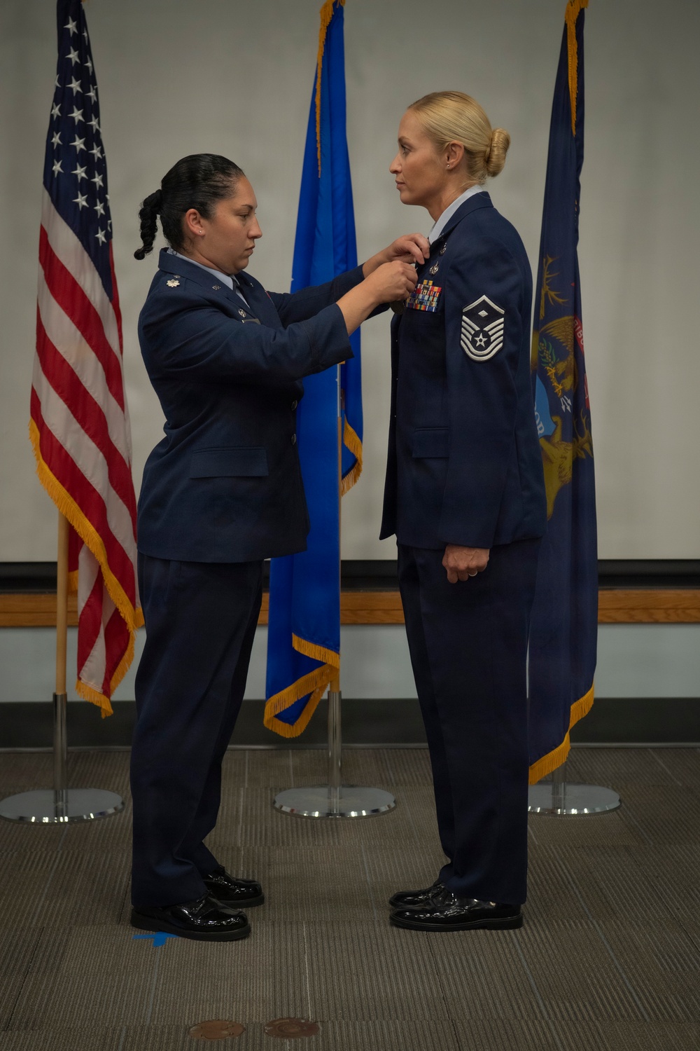 Two female U.S. Air Force personnel are participating in a formal military ceremony, with one officer pinning a medal or insignia onto the other's uniform. The recipient displays a Chief Master Sergeant rank insignia on her sleeve and multiple service ribbons on her chest. Three flags are visible in the background: the U.S. flag, an Air Force flag, and what appears to be a state flag (likely Michigan).