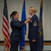 Two female U.S. Air Force personnel are participating in a formal military ceremony, with one officer pinning a medal or insignia onto the other's uniform. The recipient displays a Chief Master Sergeant rank insignia on her sleeve and multiple service ribbons on her chest. Three flags are visible in the background: the U.S. flag, an Air Force flag, and what appears to be a state flag (likely Michigan).