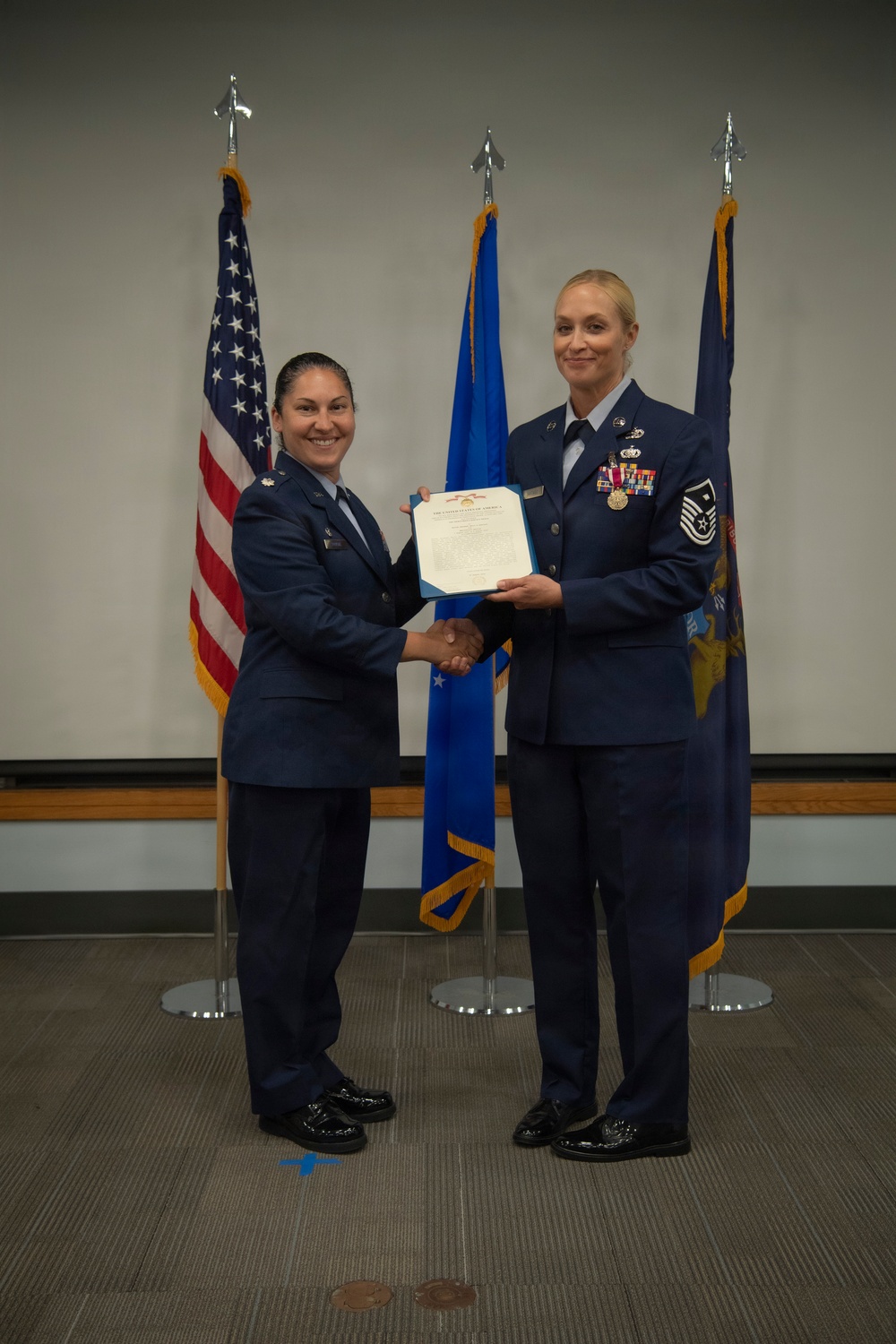 Two female U.S. Air Force personnel are posing during a formal military ceremony, shaking hands while jointly holding an official certificate or award document. The individual on the right, wearing a Senior Master Sergeant rank insignia and multiple service ribbons and medals, appears to be receiving recognition. Three flags are displayed in the background: the U.S. flag, the Air Force flag, and what appears to be a state flag.