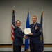 Two female U.S. Air Force personnel are posing during a formal military ceremony, shaking hands while jointly holding an official certificate or award document. The individual on the right, wearing a Senior Master Sergeant rank insignia and multiple service ribbons and medals, appears to be receiving recognition. Three flags are displayed in the background: the U.S. flag, the Air Force flag, and what appears to be a state flag.