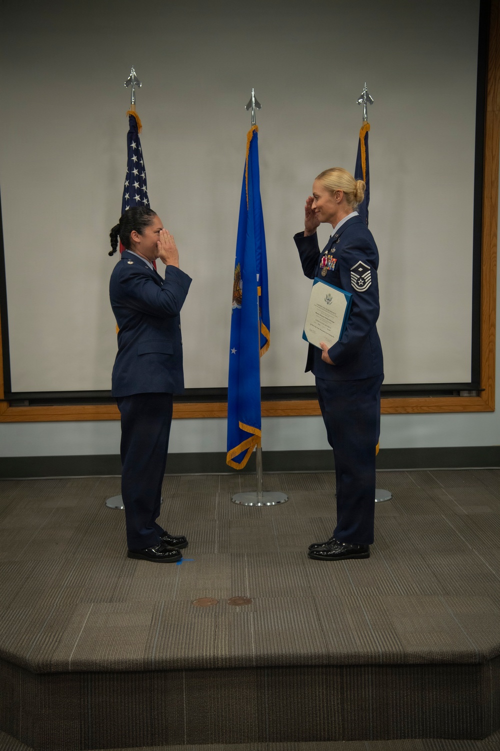 Two United States Air Force personnel in dress blue uniforms are exchanging salutes on a raised platform or stage. The individual on the right is holding what appears to be an official certificate or award document and displays multiple service ribbons/medals, indicating a formal military ceremony. Three flags are visible in the background, including the U.S. flag and what appears to be an Air Force flag.