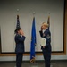 Two United States Air Force personnel in dress blue uniforms are exchanging salutes on a raised platform or stage. The individual on the right is holding what appears to be an official certificate or award document and displays multiple service ribbons/medals, indicating a formal military ceremony. Three flags are visible in the background, including the U.S. flag and what appears to be an Air Force flag.