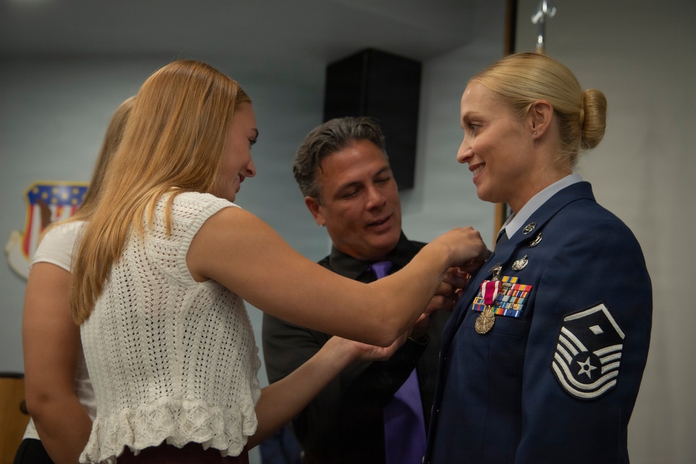 A US Air Force female senior non-commissioned officer is having a medal or insignia pinned onto her dress blue uniform during what appears to be a military promotion or award ceremony. A young blonde woman and an older man are assisting with pinning the decoration, both smiling warmly. The setting appears to be an official military facility, with a unit shield/emblem visible on the wall in the background.