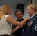A US Air Force female senior non-commissioned officer is having a medal or insignia pinned onto her dress blue uniform during what appears to be a military promotion or award ceremony. A young blonde woman and an older man are assisting with pinning the decoration, both smiling warmly. The setting appears to be an official military facility, with a unit shield/emblem visible on the wall in the background.