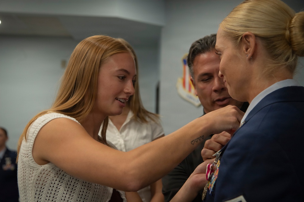 A military promotion or award ceremony is taking place indoors, likely at a U.S. Air Force installation. A young woman with long blonde hair is pinning a medal or insignia onto the uniform of a female military officer in a blue service dress uniform. A man stands closely behind, observing the pinning ceremony.