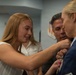 A military promotion or award ceremony is taking place indoors, likely at a U.S. Air Force installation. A young woman with long blonde hair is pinning a medal or insignia onto the uniform of a female military officer in a blue service dress uniform. A man stands closely behind, observing the pinning ceremony.