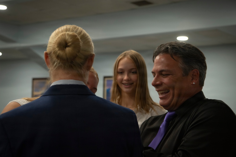A small group of people are gathered indoors in what appears to be a formal or semi-formal setting, engaged in conversation. A man wearing a black suit with a purple tie is laughing or smiling broadly, while a teenage girl with long blonde hair smiles nearby. A woman with her hair in a bun, wearing a blue blazer, is seen from behind in the foreground.