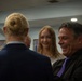 A small group of people are gathered indoors in what appears to be a formal or semi-formal setting, engaged in conversation. A man wearing a black suit with a purple tie is laughing or smiling broadly, while a teenage girl with long blonde hair smiles nearby. A woman with her hair in a bun, wearing a blue blazer, is seen from behind in the foreground.