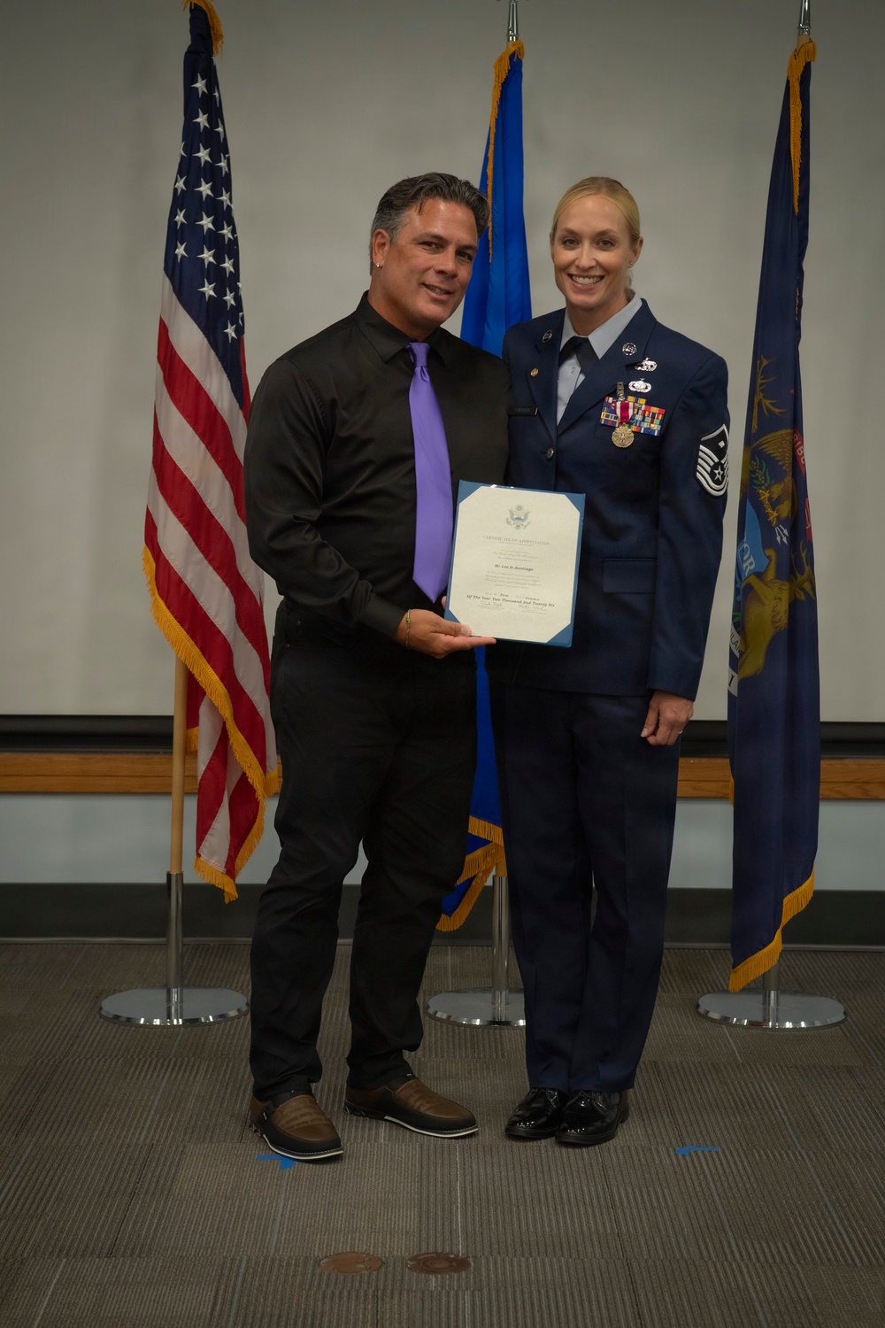 Two individuals pose together holding a certificate during what appears to be a military recognition ceremony. The woman on the right is wearing a United States Air Force dress uniform with senior NCO chevrons, multiple ribbons, and a medal. They are standing in front of three flags including the American flag and what appears to be a state flag.