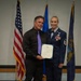 Two individuals pose together holding a certificate during what appears to be a military recognition ceremony. The woman on the right is wearing a United States Air Force dress uniform with senior NCO chevrons, multiple ribbons, and a medal. They are standing in front of three flags including the American flag and what appears to be a state flag.
