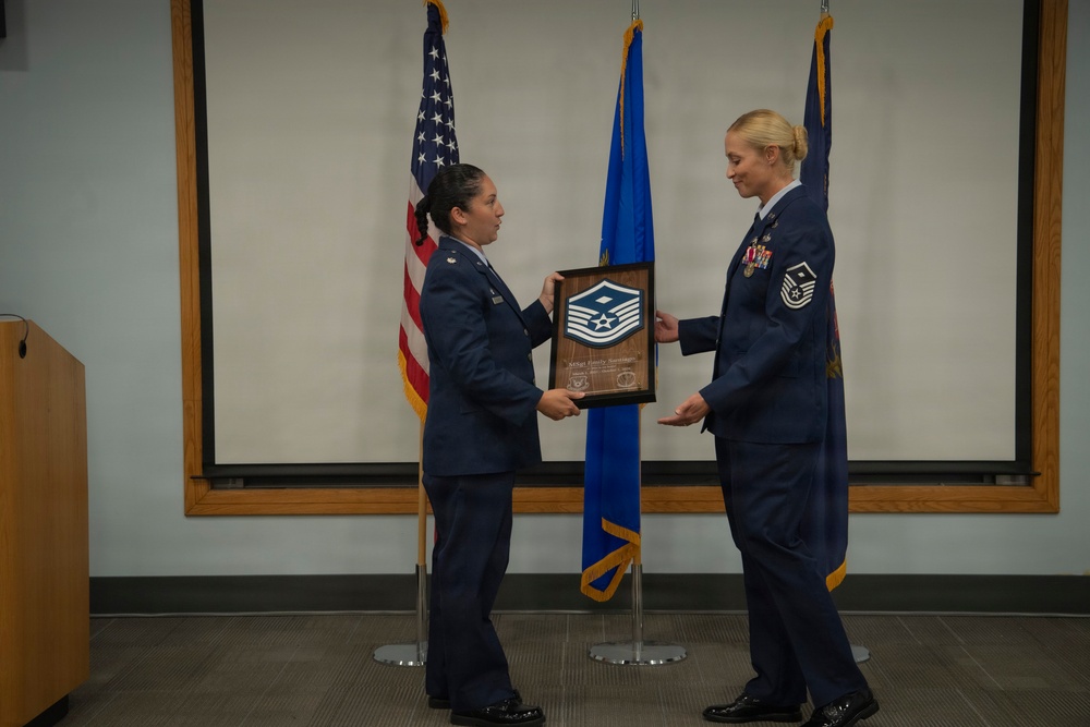 Two female U.S. Air Force personnel are participating in a formal military ceremony, with one officer presenting a wooden plaque to a Senior Master Sergeant. The plaque features the Senior Master Sergeant rank insignia and appears to be a promotion or recognition award. The ceremony takes place in front of U.S. and Air Force flags in what appears to be a conference or briefing room.